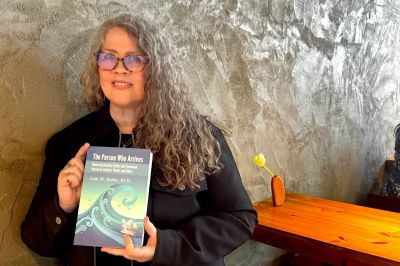 Woman with curly gray hair holding a book titled 'The Poems Who Arrives'.