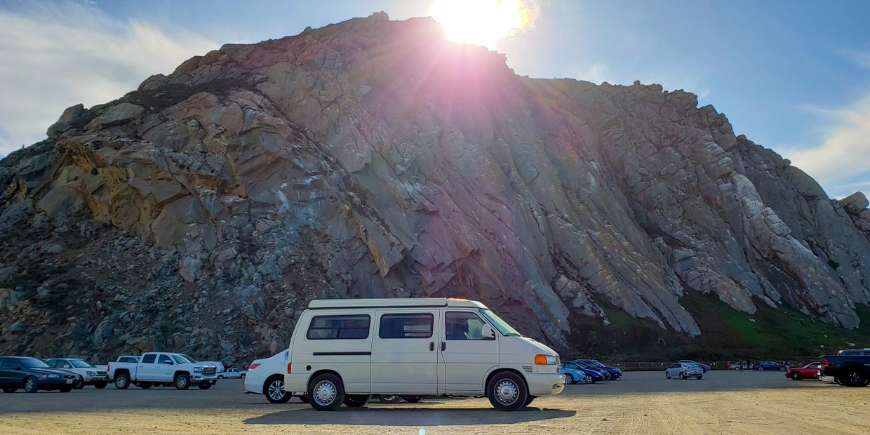 A white van parked in front of a large rocky mountain with the sun shining above.