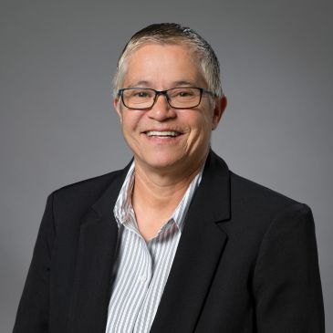 Smiling person in glasses wearing a black blazer and striped shirt against grey background.