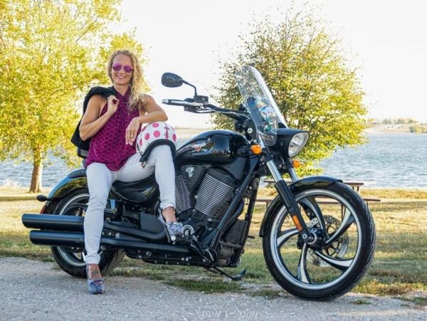A woman poses confidently on a black motorcycle by a lakeside on a sunny day.