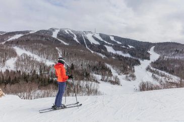 Skier in orange jacket standing on snowy slope, looking at ski trails on mountain.
