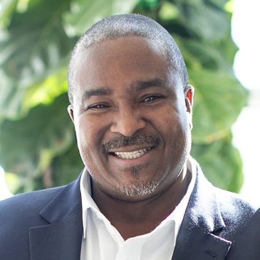 Smiling man in a dark blazer and white shirt with a leafy background.