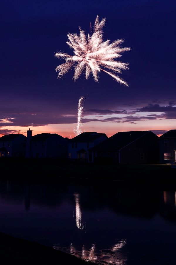 Fireworks at dusk exploding over homes with its reflection in a pond.