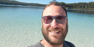 Man smiling with sunglasses at a crystal-clear lake under a blue sky.
