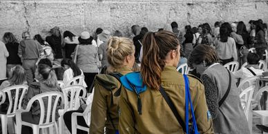 Two female soldiers in uniform stand at a prayer wall with a crowd behind them.