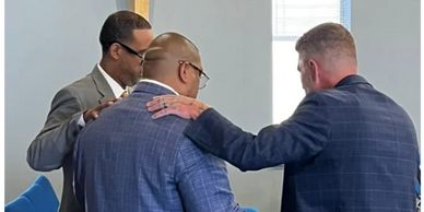 Three men in suits share a moment of prayer or support in a church setting.
