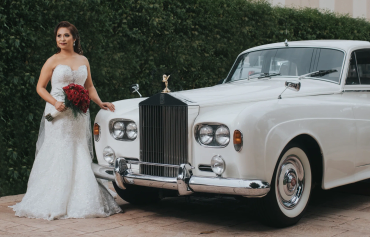 A bride in a white gown stands beside a classic white Rolls-Royce car.