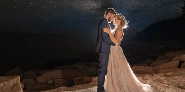 A couple in wedding attire embracing under a starry night sky on rocky terrain.