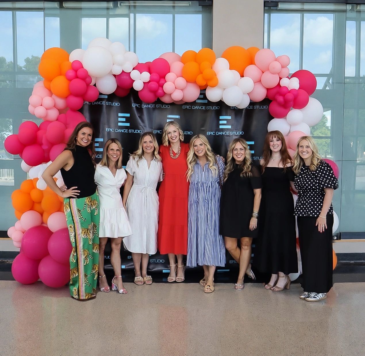 Group of eight women posing in front of colorful balloon arch.