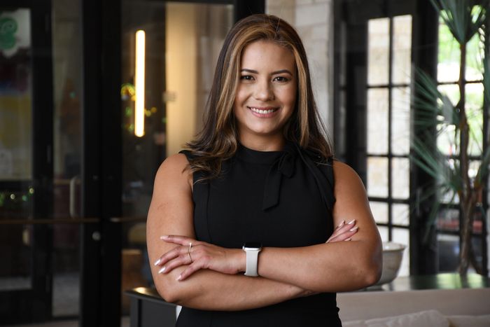 Confident woman in black dress with arms crossed, smiling at camera indoors.