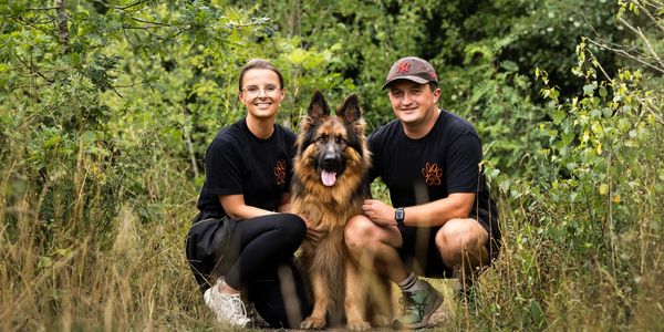 A smiling couple poses with their German Shepherd in a forest.