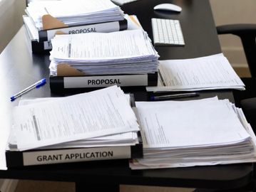 Desk cluttered with stacks of proposal documents and office supplies.