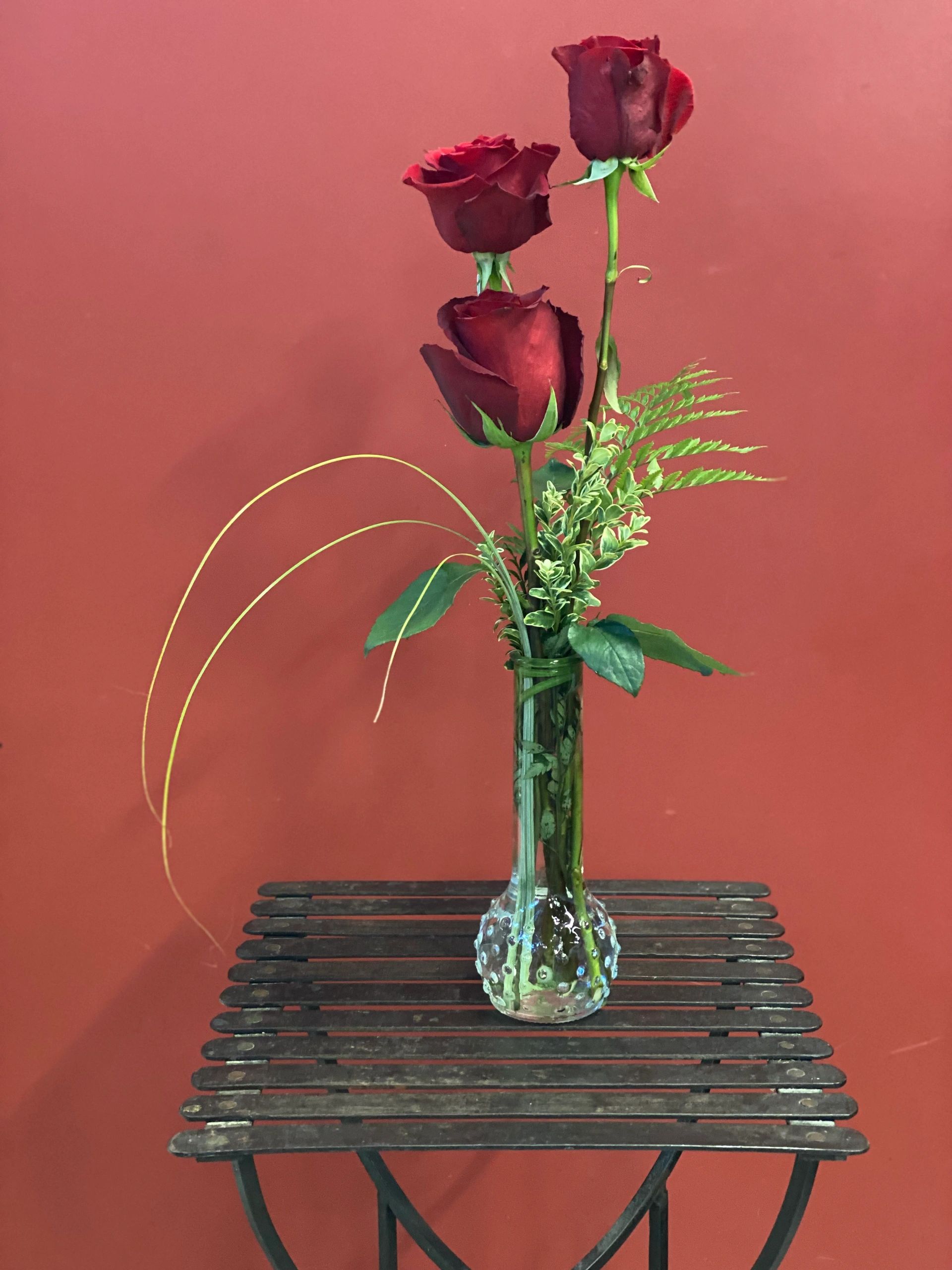 Three red roses in a decorative glass vase on a wooden table against a red wall.