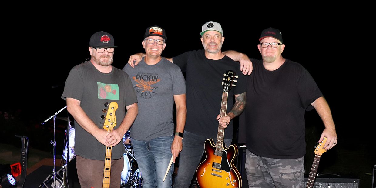 Four musicians posing with their guitars and drumsticks on stage at night.