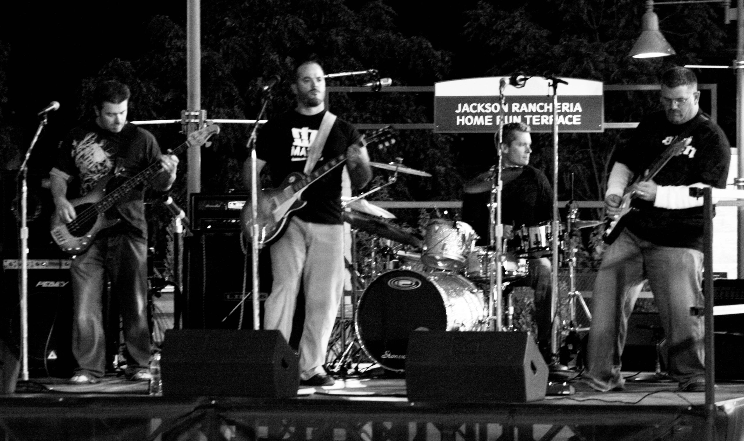 Black and white photo of a band performing on stage with guitars and drums.