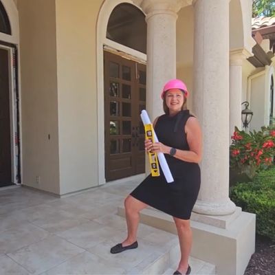 Woman in a pink hard hat holding blueprints and a level on a home's porch.