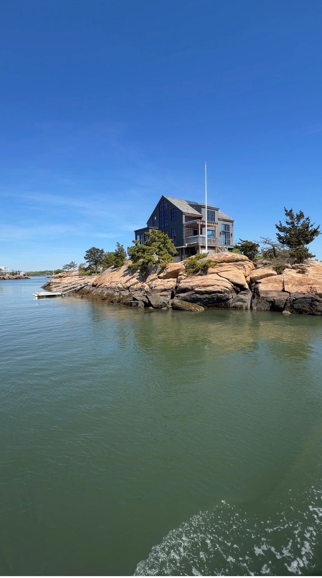 A modern house perched on rocky island surrounded by calm water under clear blue sky.
