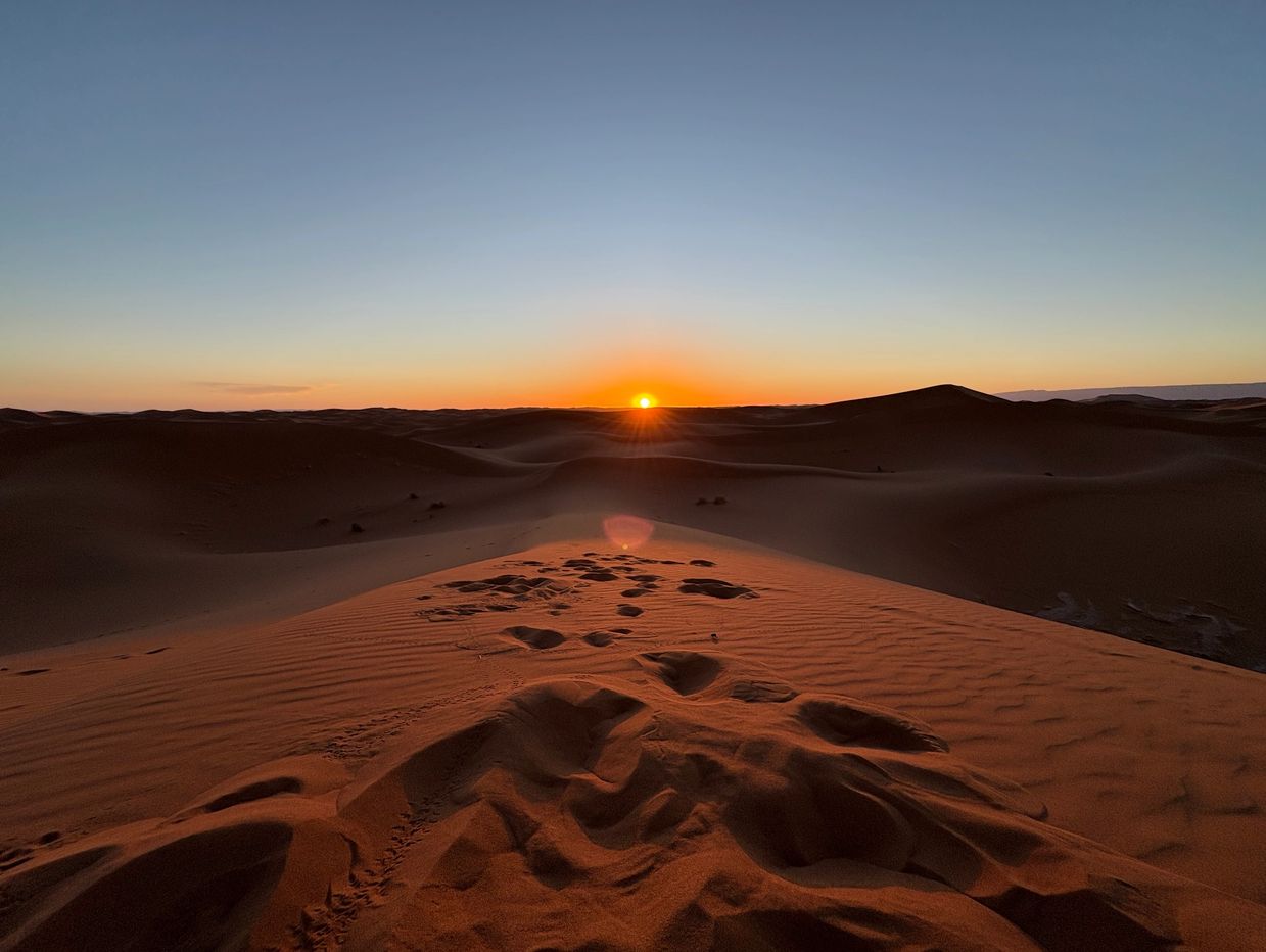Sun setting over vast desert dunes with footprints on sand.
