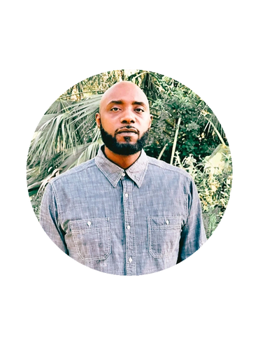 Man with a beard wearing a gray button-up shirt in a leafy outdoor setting.