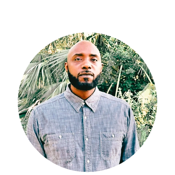 Man with a beard wearing a gray button-up shirt in a leafy outdoor setting.