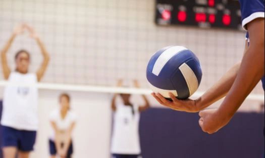 Player preparing to serve a volleyball in a gym with teammates in the background.