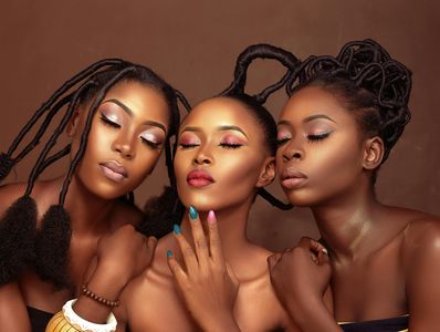 Three women with artistic braided hairstyles and vibrant makeup pose with closed eyes.
