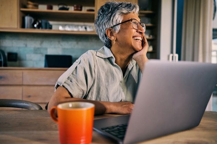 Smiling elderly woman working on laptop with orange mug nearby.