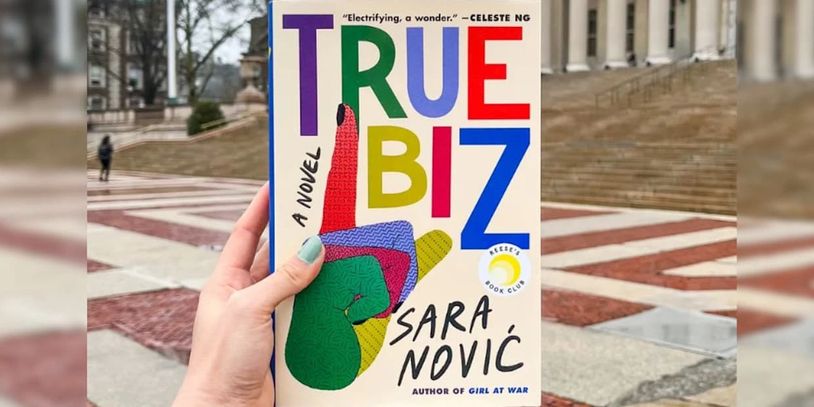 Cover of book, True Biz by Sara Novic held up in front of a historic staircase