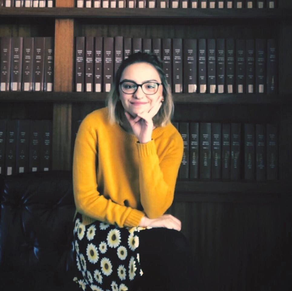 Woman in glasses and yellow sweater sitting in front of a bookshelf.