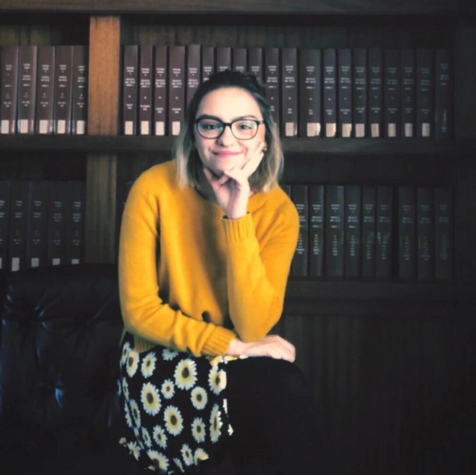 Woman in glasses and yellow sweater sitting in front of a bookshelf.