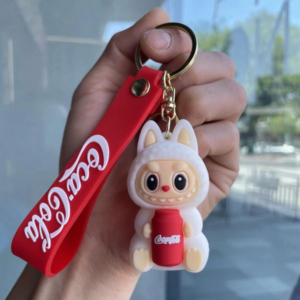 Hand holding a cute Coca-Cola keychain with a cartoon character and red strap.