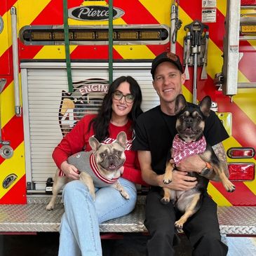 Couple with two French Bulldogs sitting on a fire truck bumper, smiling.