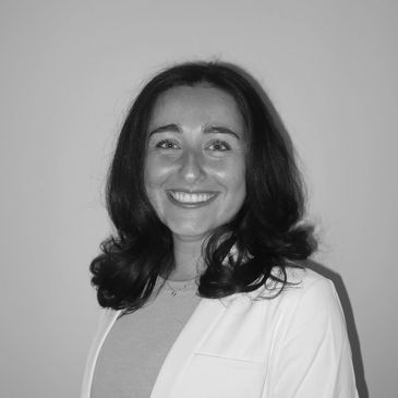 Smiling woman with dark hair wearing a white blazer against a plain background.