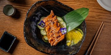 Grilled salmon with sesame seeds, cucumber, and edible flowers on a black plate.
