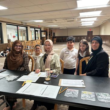 Group of women and children sitting and standing at a table with papers in a community hall.