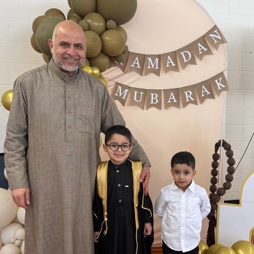 A man with two boys posing in front of a Ramadan Mubarak banner.
