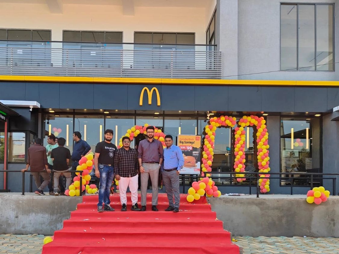 Four men standing on a red carpeted staircase outside a McDonald's with balloon decorations.