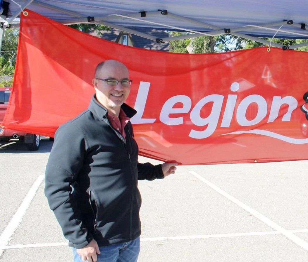 Man smiling and holding a red Legion banner under a canopy outdoors.