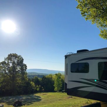 RV parked on a sunny hillside with trees and distant hills.
