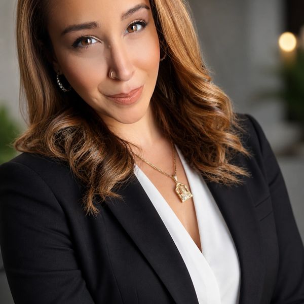 Confident woman with wavy hair wearing a black blazer and gold necklace.