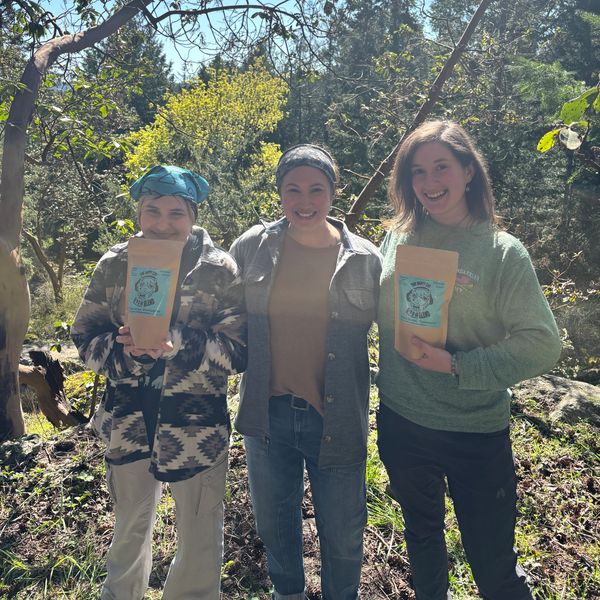 Three people outdoors holding bags of coffee, smiling in sunlight.