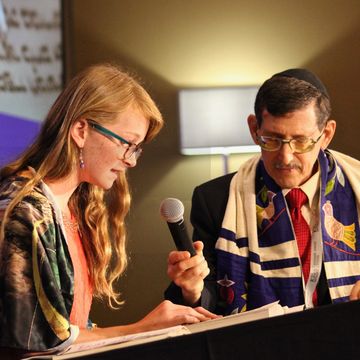 A young woman and a man with a tallit and kippah reading together at a table.
