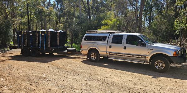 Silver pickup truck towing a trailer loaded with secured cargo in a wooded area.