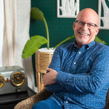 Smiling bald man with glasses sitting on a chair.