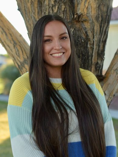 Smiling woman with long dark hair standing by a tree outdoors.