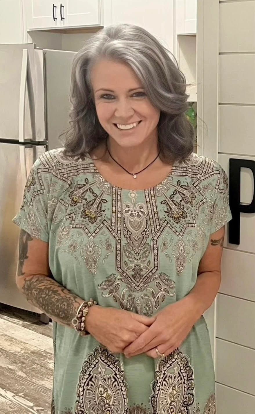 Smiling woman with gray hair and tattoos in a patterned dress.