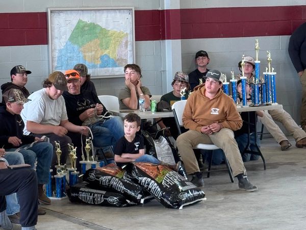 Group of people, mostly boys, sitting indoors with trophies and bags around.