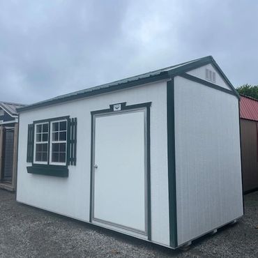 White storage shed with green trim and window shutters on gravel.