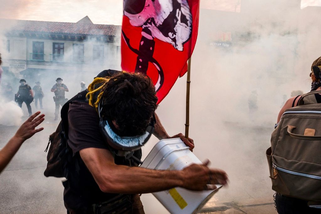 Man running towards camera, away from tear gas canister. Man is holding a red flag and a bucket.