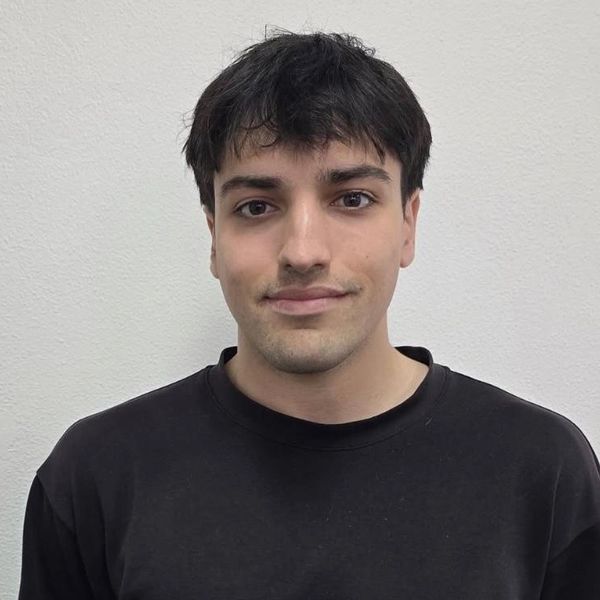 Young man with dark hair wearing a black shirt against a plain wall.